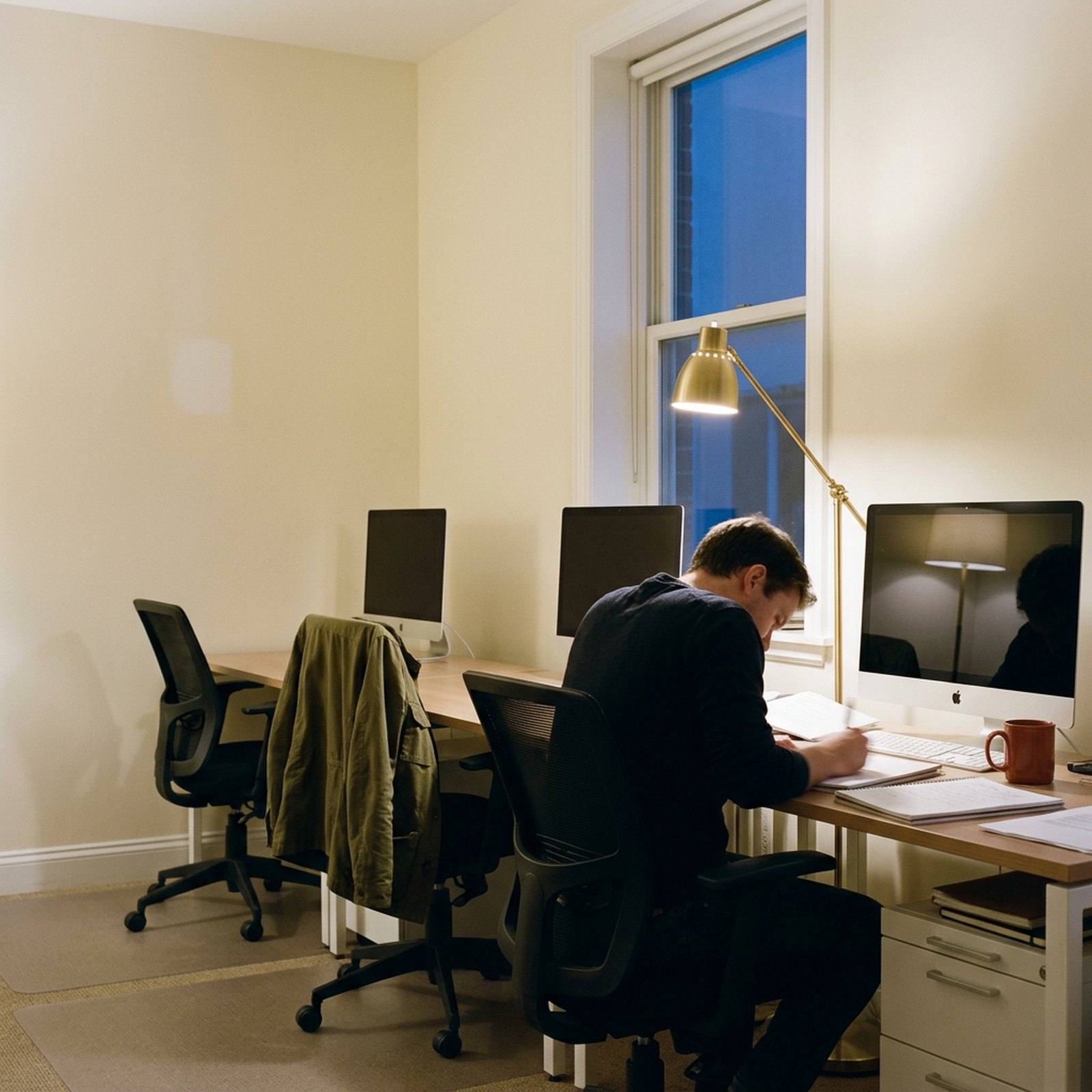 Un open space en fin de journée avec deux bureaux vides et un troisième occupé de dos, une veste olive sur le dossier d'une chaise