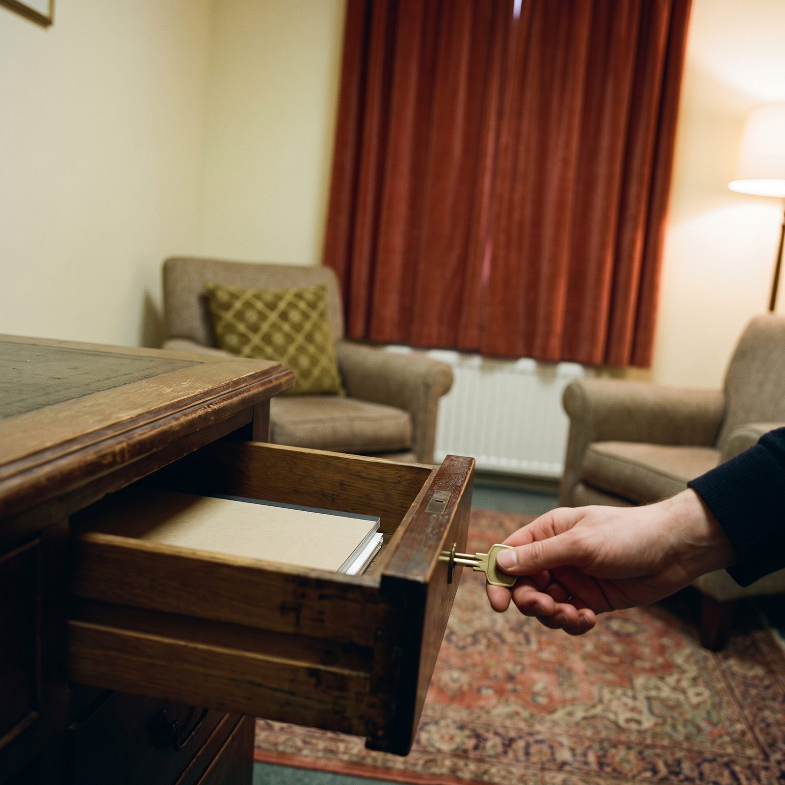 Une main referme à clé le tiroir en bois d'un bureau de thérapeute, deux fauteuils face à face en arrière-plan, aucun ordinateur dans la pièce
